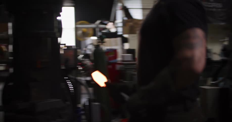 A tattooed blacksmith pulls a glowing hot piece of metal from a large forge and carefully places it into a smaller one across the workshop