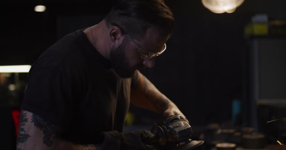 In slow motion, a bearded and tattooed artisan in safety glasses grinds a piece of metal, creating a brilliant shower of sparks in his workshop