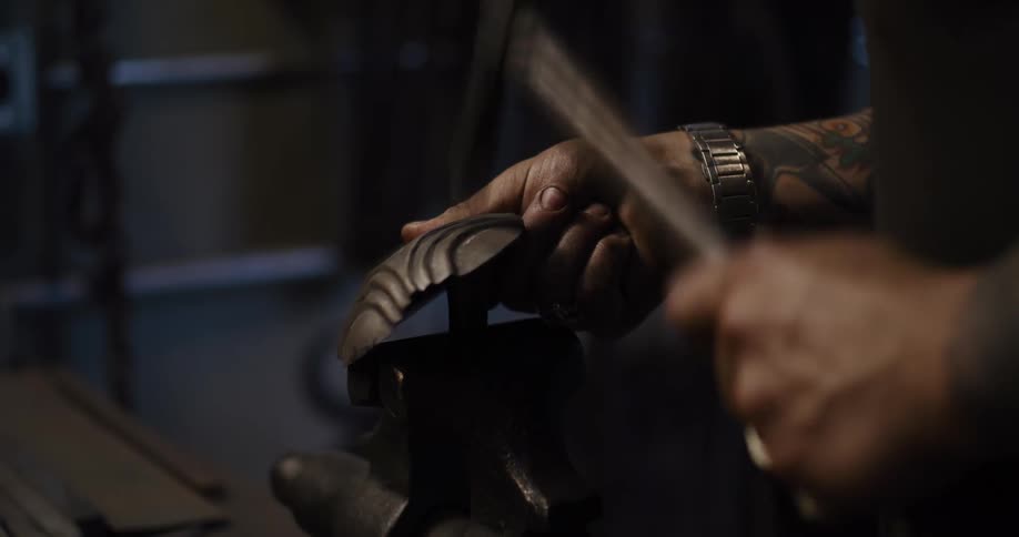 A tattooed metalworker hammers and shapes a piece of metal on a vise in a dark workshop