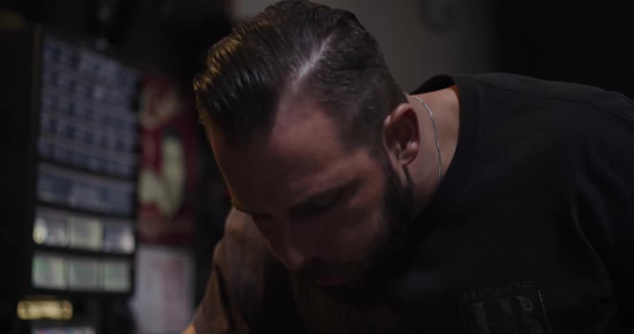 A tattooed craftsman carefully sands a piece of wood held in a vise in his workshop, shown in slow motion