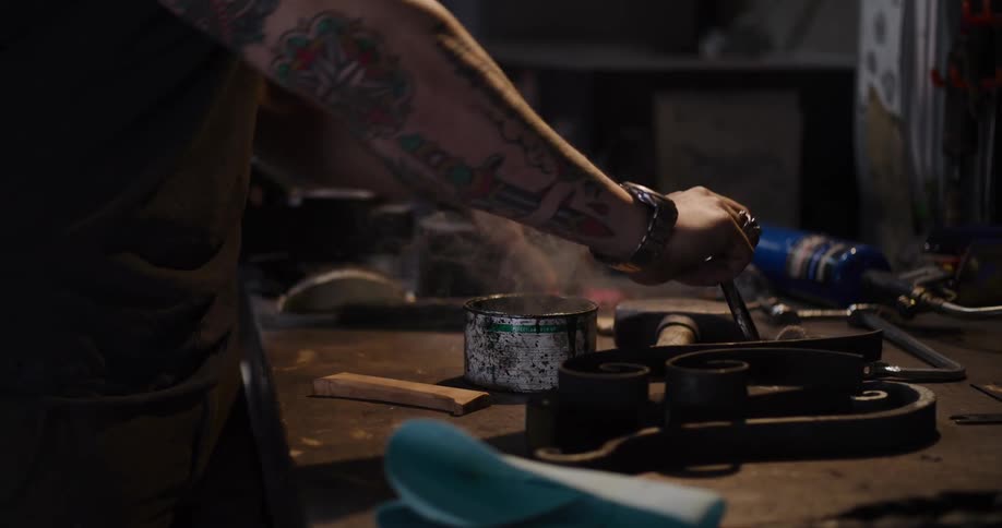 A tattooed artisan uses a blowtorch to light a fire in a can, then uses the contents to paint a piece of metal in his workshop