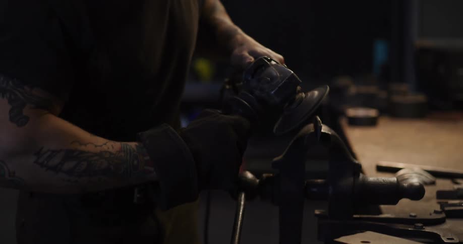 A tattooed man in a dark workshop grinds metal with an angle grinder, creating a spectacular shower of sparks in slow motion