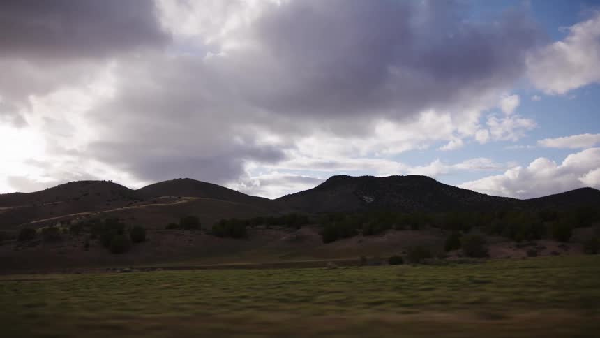 A scenic drive through a vast countryside of rolling green hills under a dramatic, partly cloudy sky