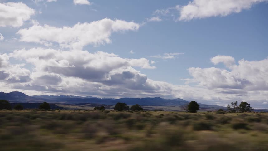 Tracking shot from a moving vehicle captures the vast, open plains and distant mountains of the American West under a cloudy sky