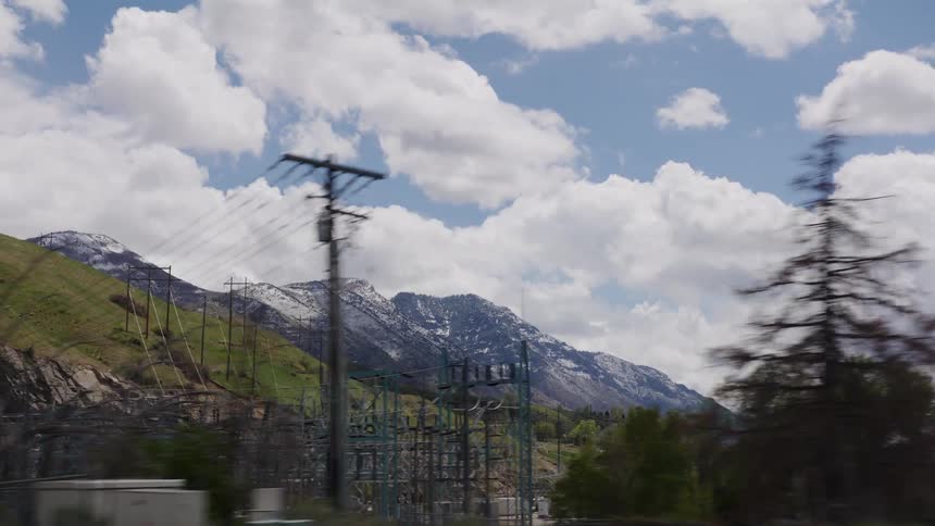 A slow-motion drive-by shot reveals a sprawling power substation at the base of lush green hills and snow-dusted mountains