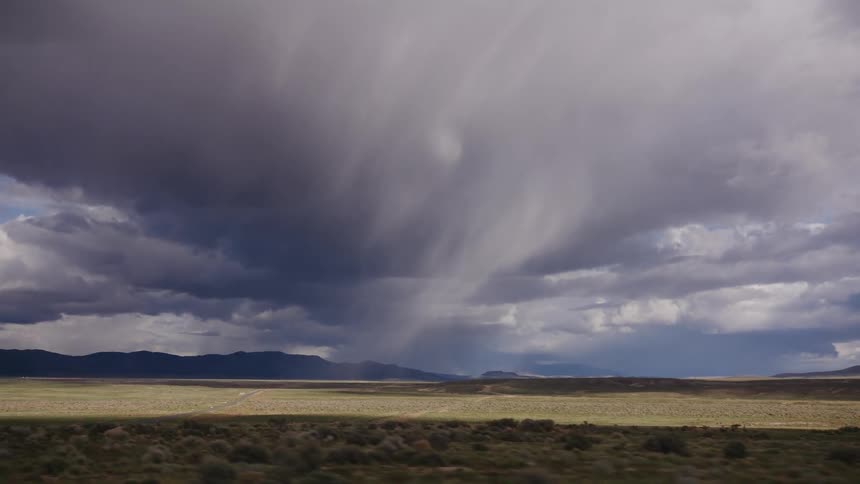 Slow motion driving shot captures a dramatic storm cloud releasing a downpour over a vast, remote landscape