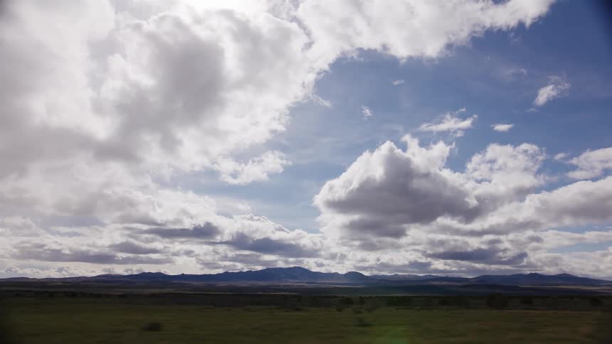 A scenic view from a moving vehicle across a vast, open plain with distant mountains under a dramatic cloudy sky