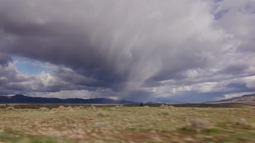 A powerful rain storm forms over a vast, arid landscape as seen from the window of a moving vehicle