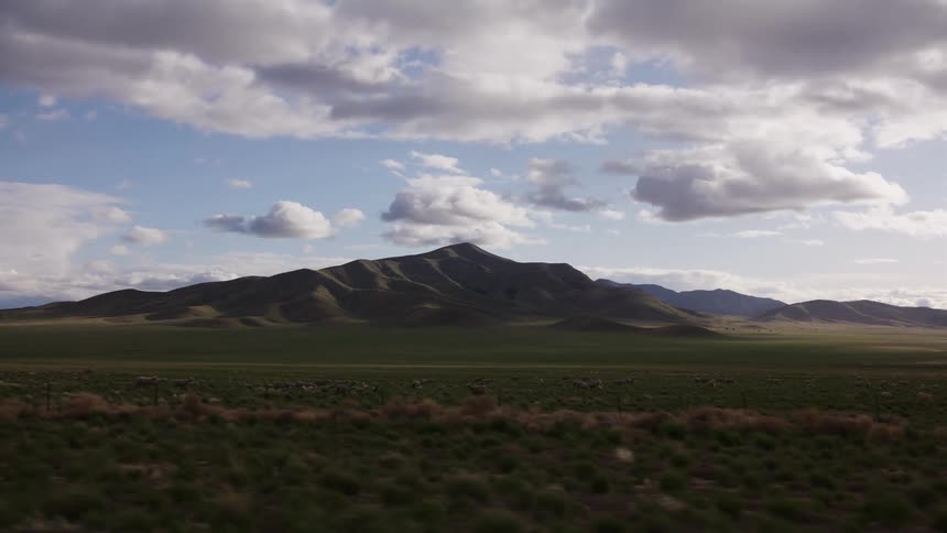 A majestic mountain range towers over a vast green valley where a herd of animals grazes under a dramatic cloudy sky