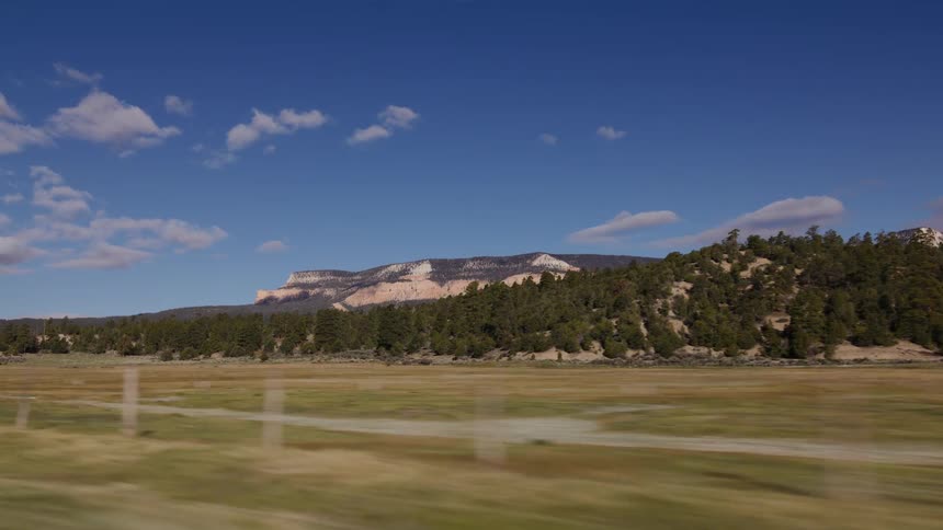 A scenic side-view from a moving vehicle reveals a vast field and forested hills with a majestic mountain range in the distance