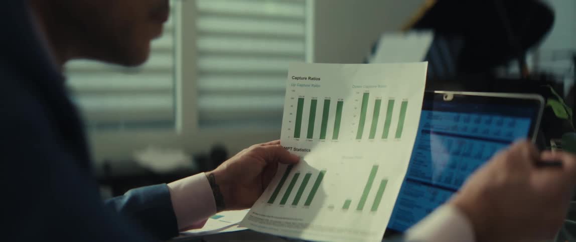 A serious businessman in glasses analyzes financial charts on paper while cross-referencing with data on his laptop in a modern office
