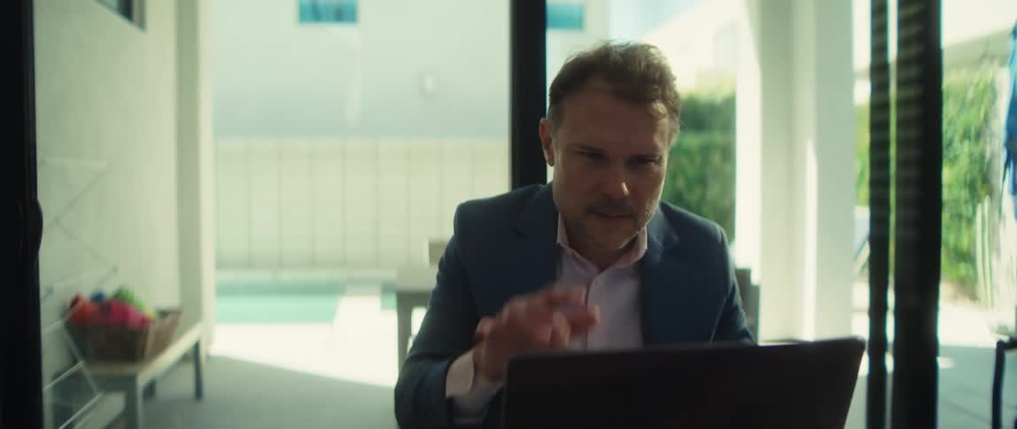 Slow zoom on a happy businessman in a suit working on his laptop from his modern home with a view of the pool