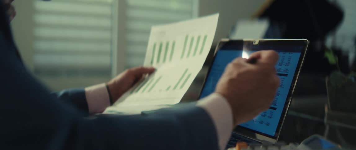 A financial analyst cross-references data from a printed report with a spreadsheet on his laptop while using a calculator