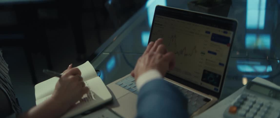 A businessman points to a financial graph on a laptop screen while his colleague takes notes during a meeting