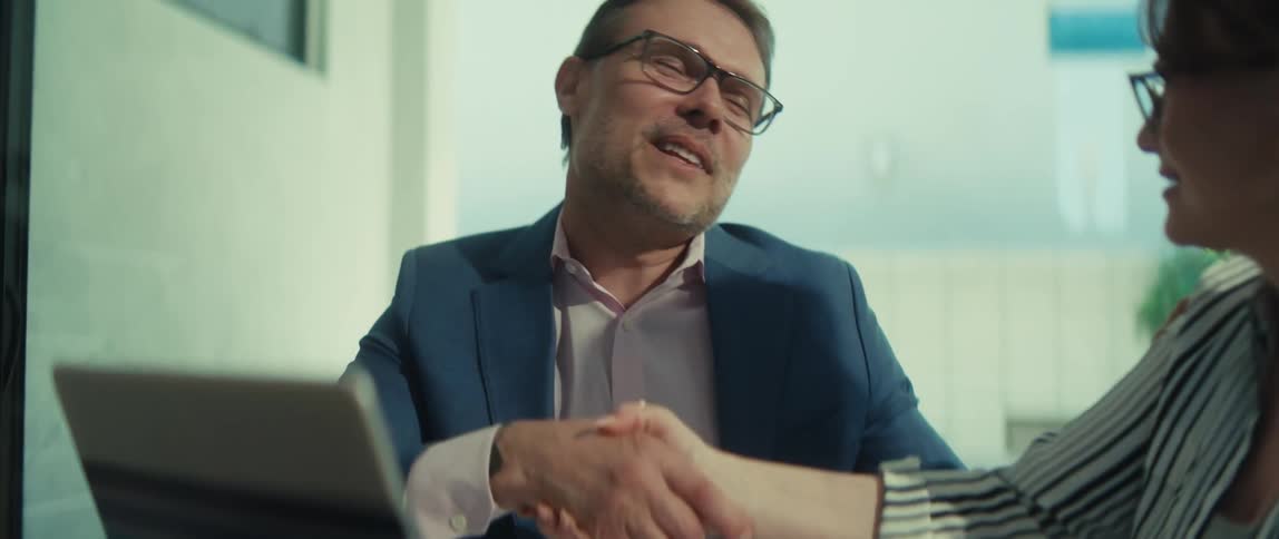 A smiling businessman and his female colleague finalize a deal with a firm handshake during a meeting in a bright, modern office
