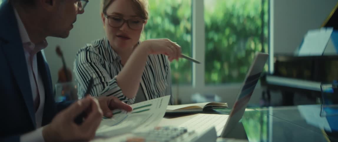 A man and a woman in business attire have a focused discussion over financial reports and a laptop in a modern office