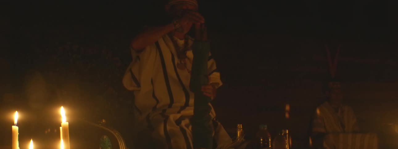A shaman in traditional attire performs a sacred candlelit ceremony, playing a wooden flute in the darkness