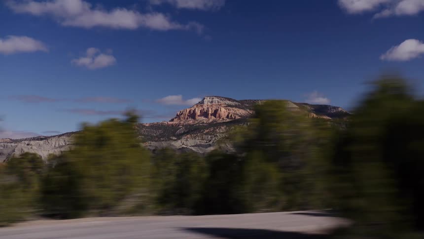 A scenic drive through a high desert landscape, with blurred trees rushing by in the foreground revealing a majestic layered mountain range in the distance