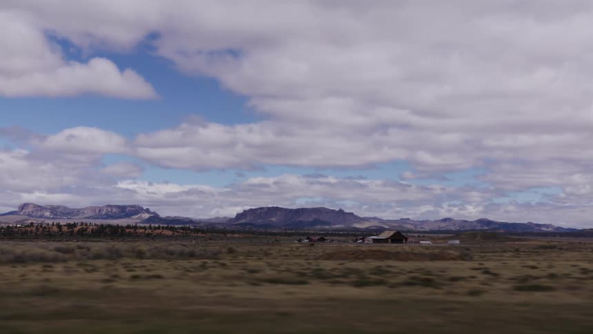A scenic drive through a vast desert landscape reveals distant mesas and a solitary barn under a partly cloudy sky