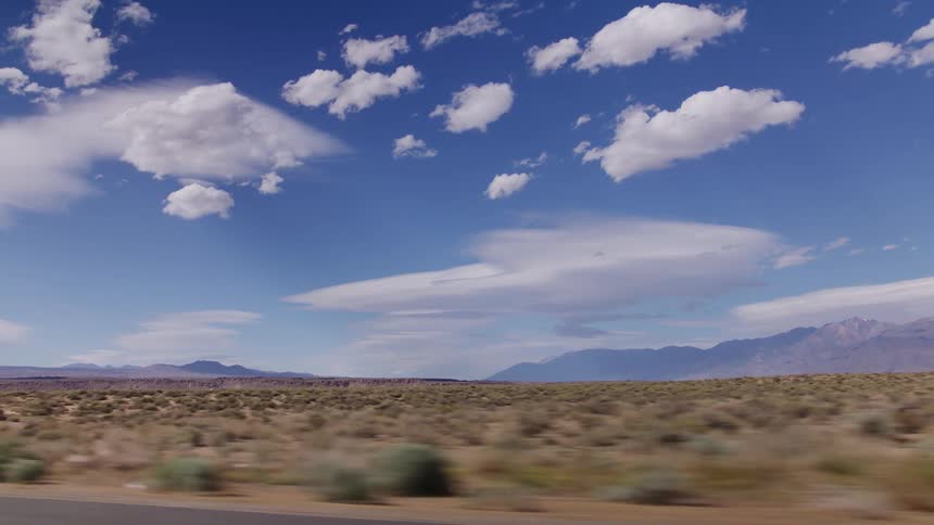 A scenic drive through a vast desert landscape revealing distant mountains under a bright blue sky with white clouds
