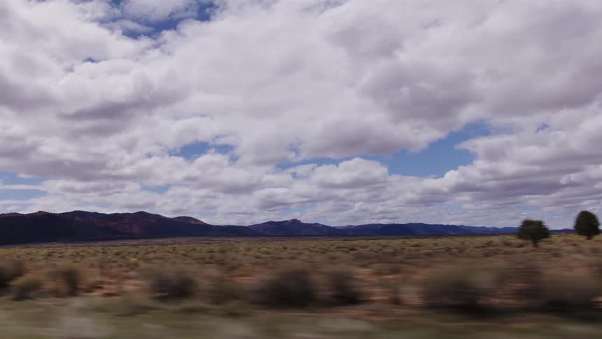 Side-view shot from a moving vehicle captures the sweeping desert landscape and distant red mountains under a wide, cloudy sky