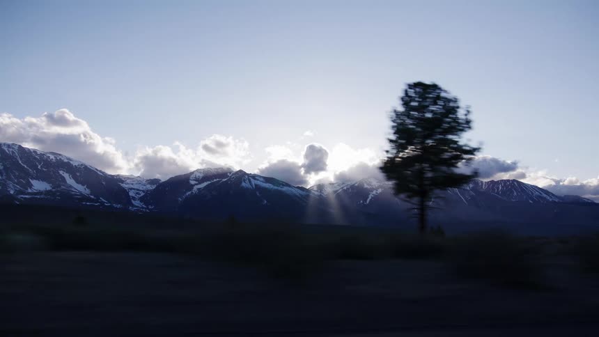 A fast-moving tracking shot reveals a majestic snow-capped mountain range as the sun shines through the clouds