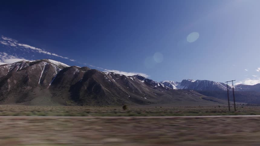 Driving alongside a vast, arid plain with majestic, snow-capped mountains looming in the distance under a sunny blue sky