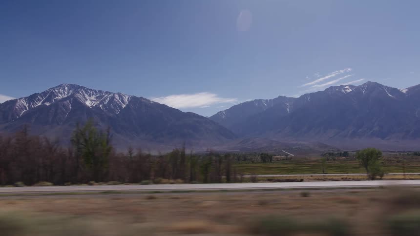 A scenic passenger-side view of a majestic snow-capped mountain range while driving through a vast valley on a sunny day