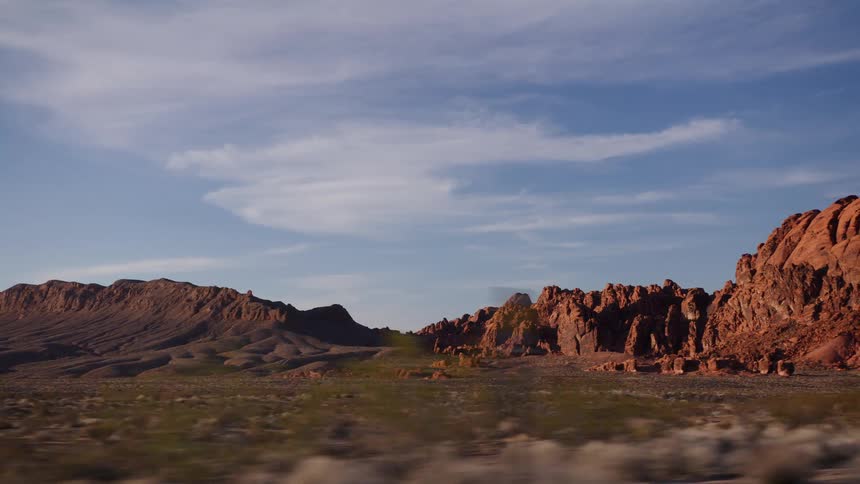 A scenic driving shot passes majestic red rock formations and mountains in a vast desert landscape under a blue sky