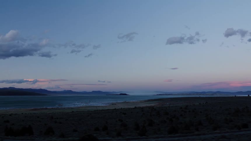 Tracking shot reveals a serene view of a large lake surrounded by mountains at dusk, under a soft pastel sky