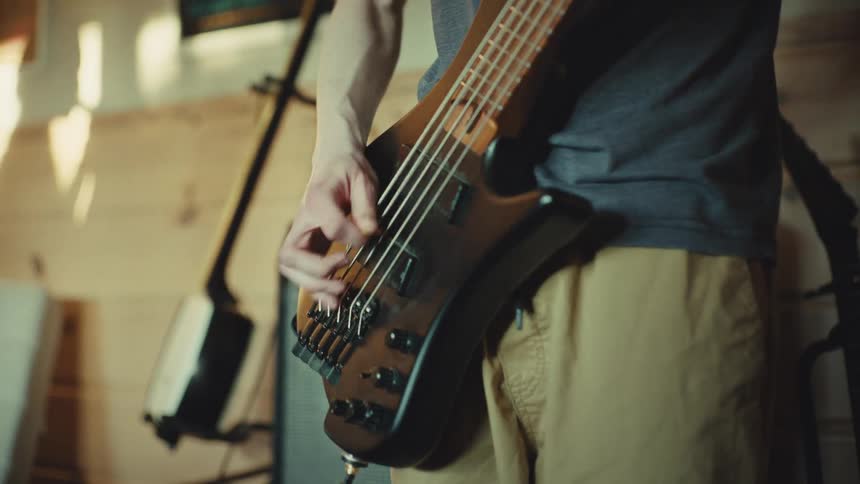 Close-up of a musician's hands as they energetically strum a five-string electric bass guitar with a pick in a sunlit music room