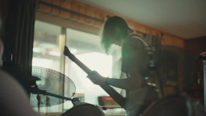 From behind a drum kit, a long-haired guitarist shreds on his electric guitar while a female singer performs during an energetic band practice