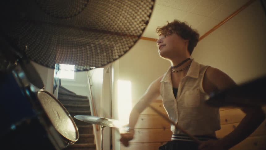 A passionate young drummer smiles while playing a groovy beat on their drum kit in a hazy, sun-drenched room