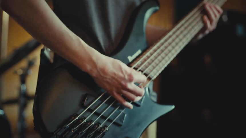 Focused young musician with medium-length hair plays a fast riff on his five-string electric bass guitar in a practice room