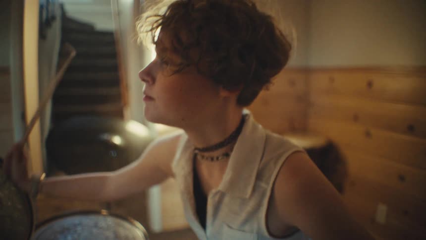 A smiling teenager with braces and short hair joyfully plays the drums in a sunlit basement practice room