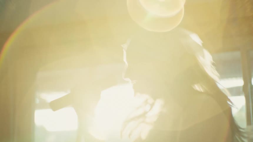 A passionate young woman in a bandana sings into a microphone, her silhouette backlit by the bright, hazy sun creating a beautiful lens flare