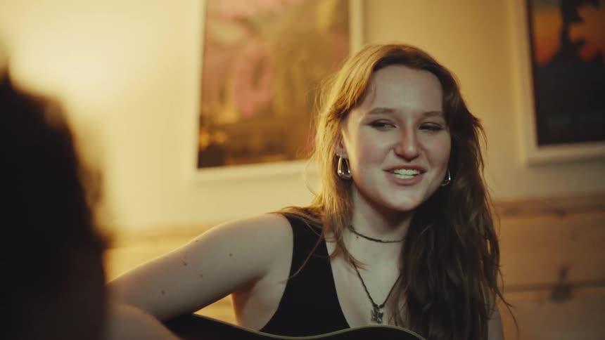 Joyful young female musician sings and plays an acoustic guitar for a friend in a warmly lit, cozy room