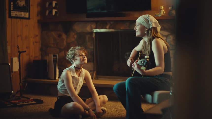 A young woman plays a song on her acoustic guitar for her friend as they sing together in a cozy, rustic room