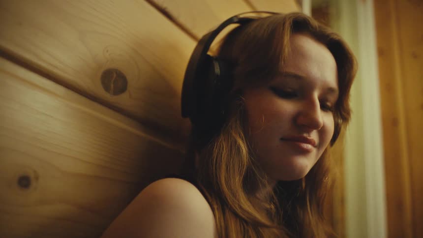 A serene young woman with headphones on leans against a wooden wall, lost in the music she's listening to