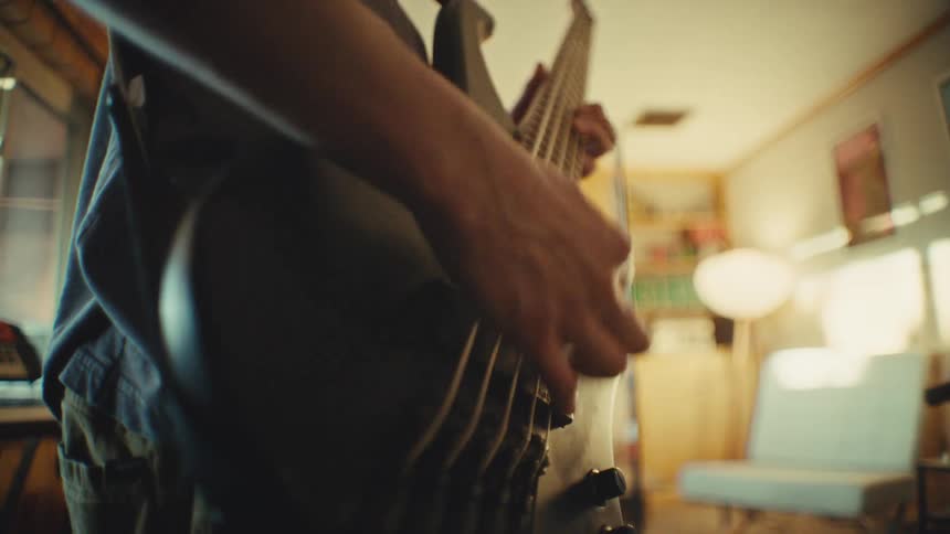 Low-angle slow-motion shot of a bassist playing an electric bass guitar during a band rehearsal in a sunlit room