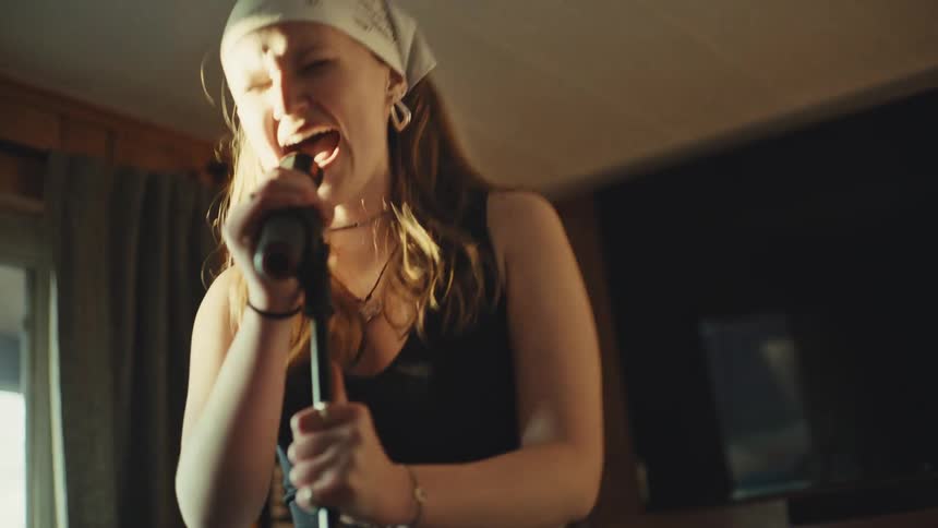 A female singer wearing a white bandana passionately sings into a microphone in slow motion while headbanging in a sunlit room