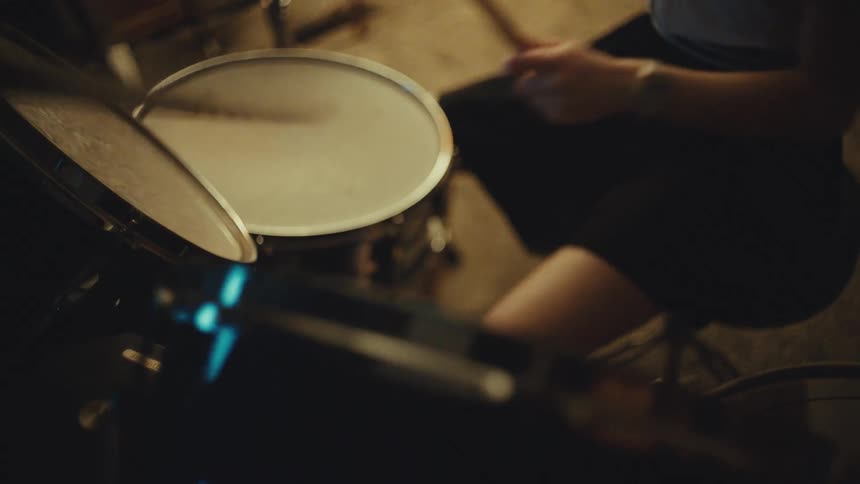A young female drummer energetically plays a beat on her drum kit in a warmly lit, wood-paneled room