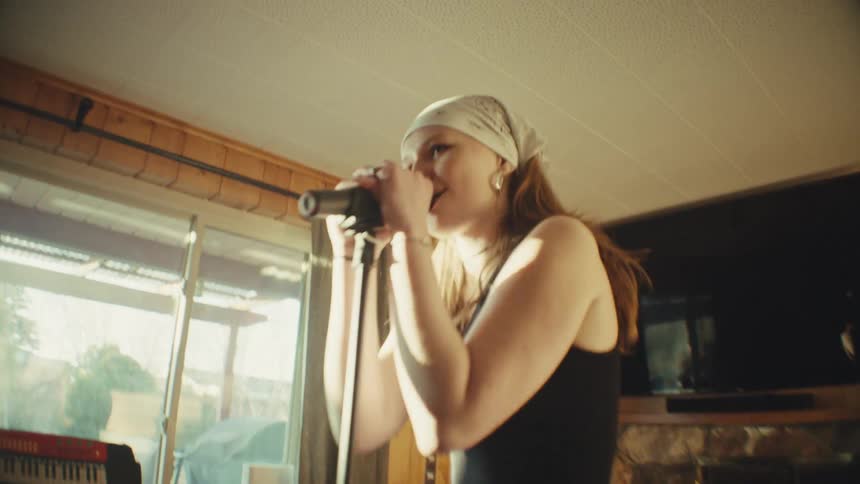 A passionate young woman wearing a bandana sings into a microphone during a sunlit band rehearsal at home