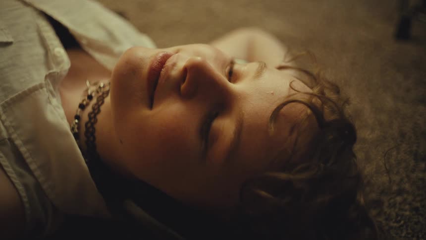 A thoughtful young woman with curly hair closes her eyes peacefully while lying on a carpeted floor in a warmly lit room
