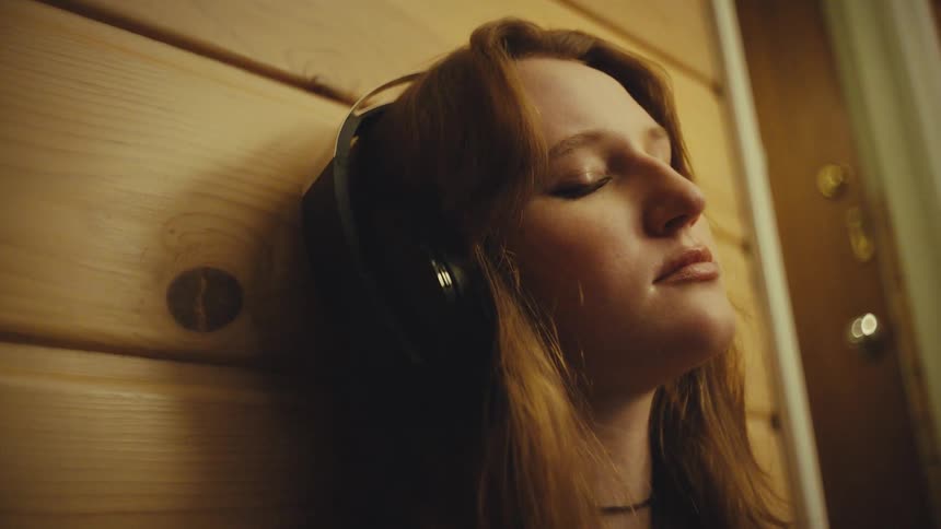 Close-up of a serene young woman with eyes closed, relaxing against a wooden wall while listening to music through headphones in a warmly lit room