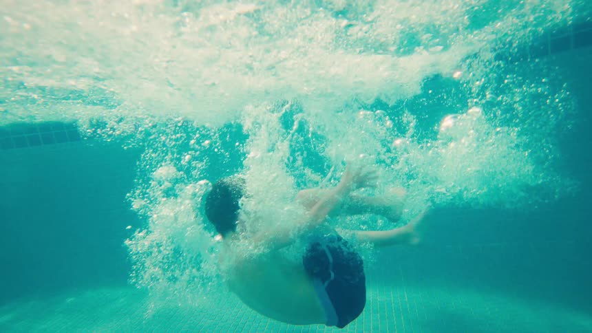 A young boy cannonballs into a tropical resort pool in dramatic slow motion, filmed from a dynamic half-submerged perspective