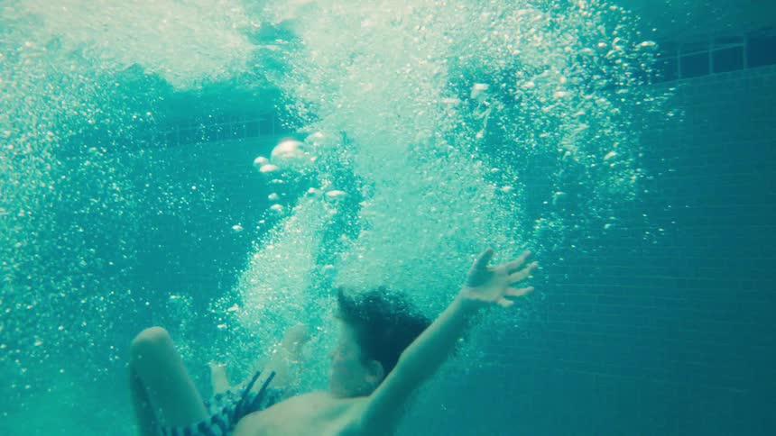 A young boy cannonballs into a swimming pool in slow motion, shot from the water's surface and then from underwater