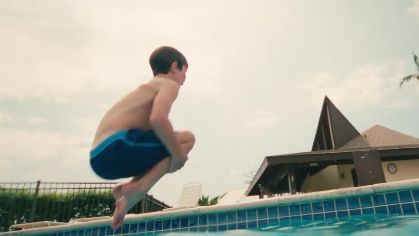 A young boy takes a running leap into a swimming pool in slow motion at a sunny tropical resort