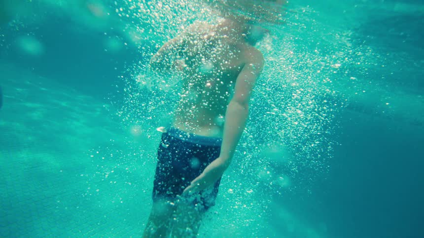 Slow motion underwater shot of a young boy gracefully moving through sunlit, bubbly water in a swimming pool