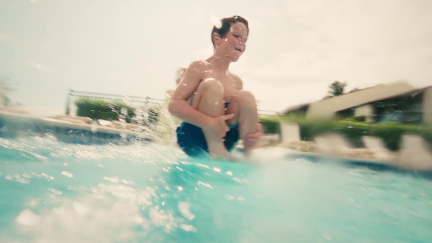 A young boy takes a running leap and cannonballs into a swimming pool, filmed in dynamic slow motion from the water's surface
